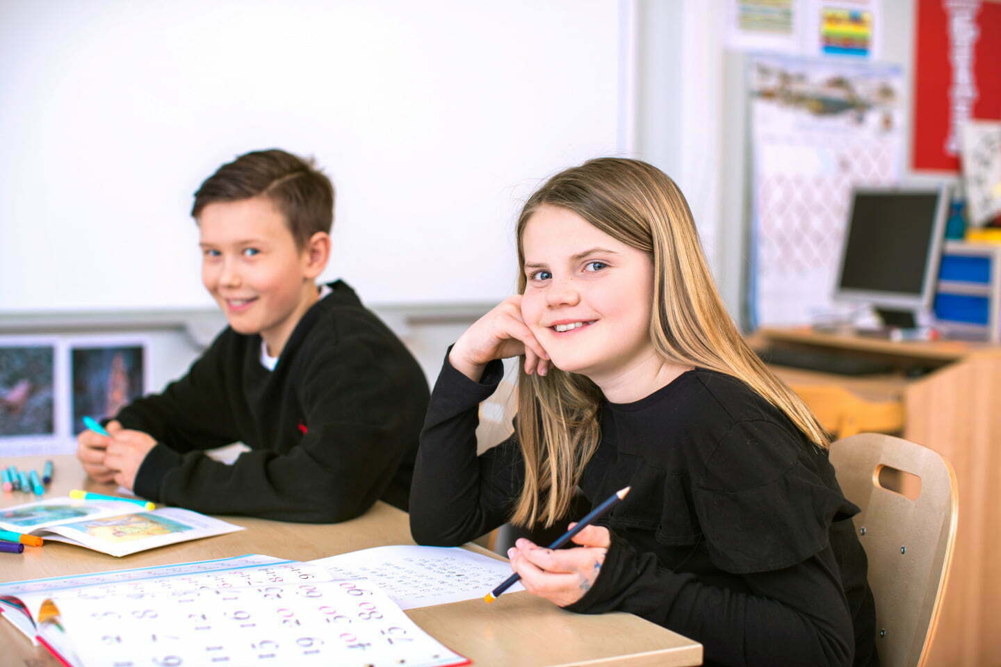 pojke och flicka, båda i svart tröjor sitter vid skolbänk, tittar in i kamera