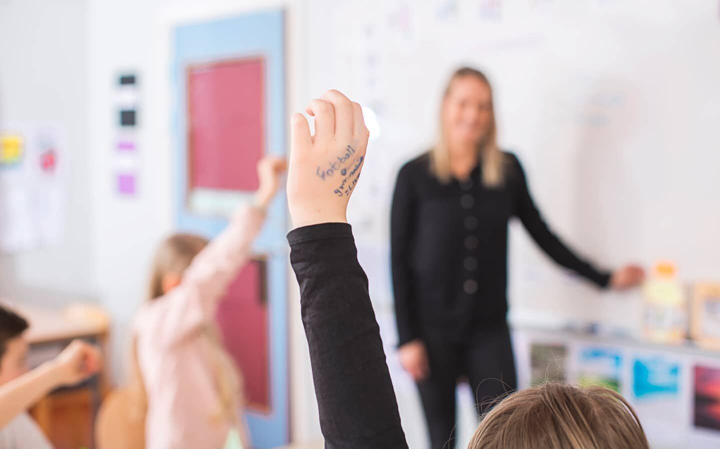 Barn som räcker upp handen framför lärare. Uppsträckt hand i fokus
