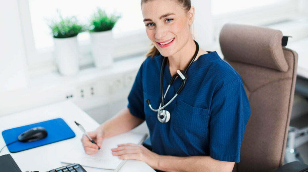 kvinna i blått med stetoskop runt halsen vid skrivbord